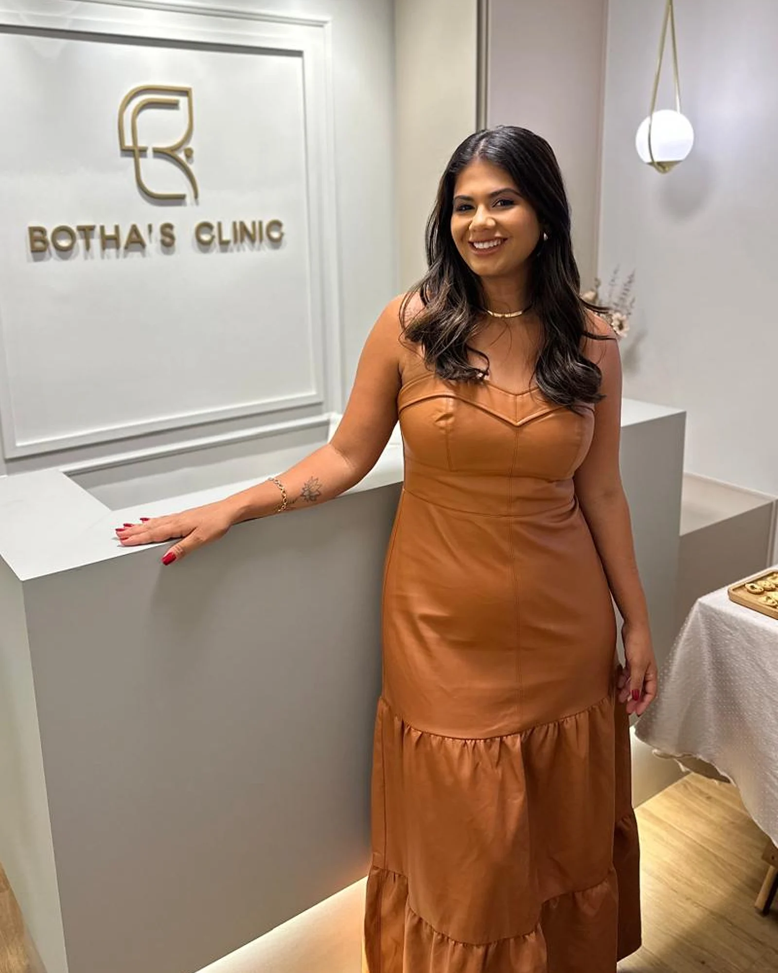 Dra. Ávila Botha sorrindo na recepção da Botha's Clinic, diante do letreiro dourado